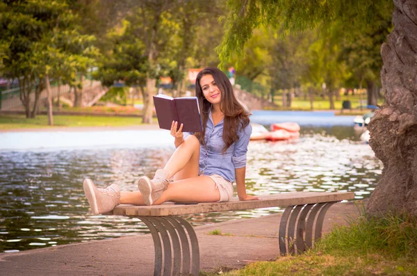 Brunette model wearing denim shirt and white shorts relaxing in park environment, sitting on bench next to lake reading book