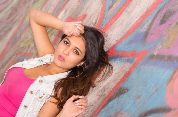 Closeup brunette model wearing pink top, white vest lying down on concrete skatepark surface with graffiti visible, looking at camera