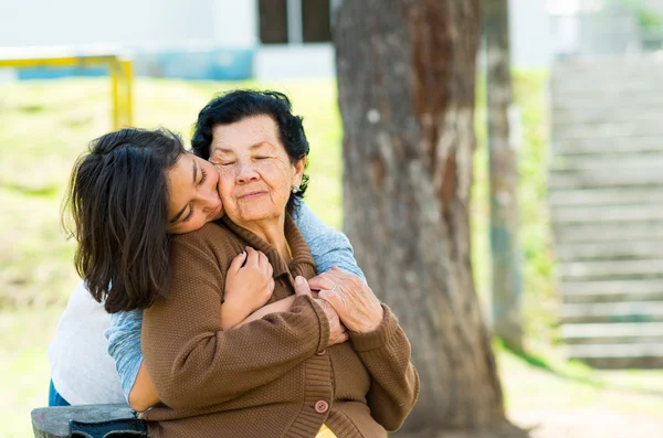 Young girl standing behind grandmother hugging and embracing in outdoors environment