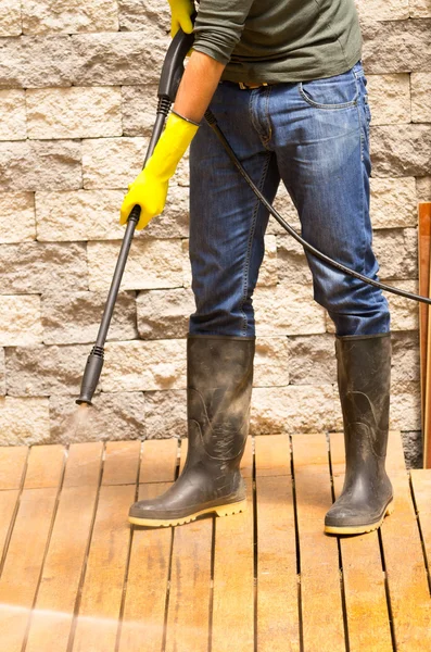 Man wearing rubber boots using high water pressure cleaner on wooden terrace surface