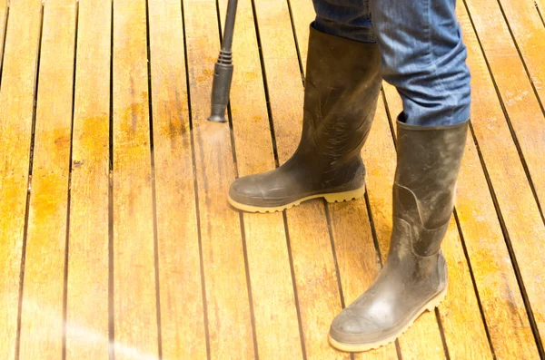 Man wearing rubber boots using high water pressure cleaner on wooden terrace surface