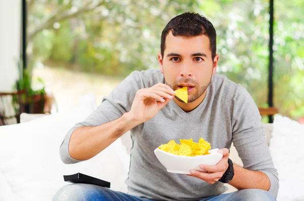 Hispanic male wearing light blue sweater plus denim jeans sitting in white sofa holding bowl of potato chips and remote control watching tv enthusiastically