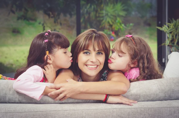 Hispanic mother in sofa with two daughters kissing her cheeks from each side, blurry garden background