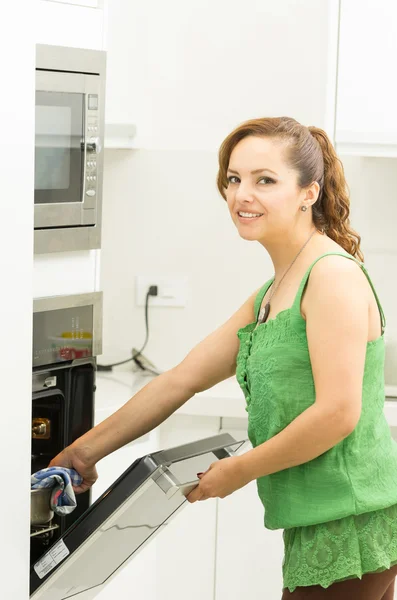Woman wearing green top in modern kitchen holding mittens and opening oven door