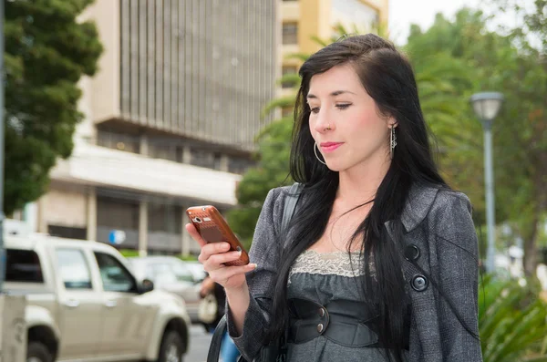 Classy latina model wearing smart casual clothes walking in urban street looking at mobile phone screen