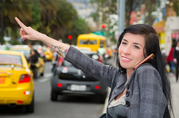 Classy latina model wearing smart casual clothes walking in urban street holding arm out signalling for taxi while talking on phone