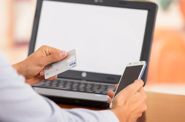 Closeup of young mans hands holding smartphone up, Visa card in other hand with laptop computer sitting on desk