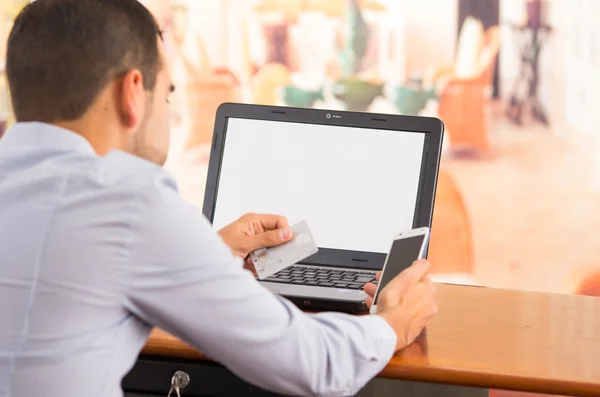 Closeup of young mans hands holding smartphone up, Visa card in other hand with laptop computer sitting on desk