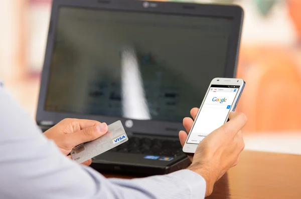 Closeup of young mans hands holding smartphone up with Google website visible, Visa card in other hand with laptop computer sitting on desk