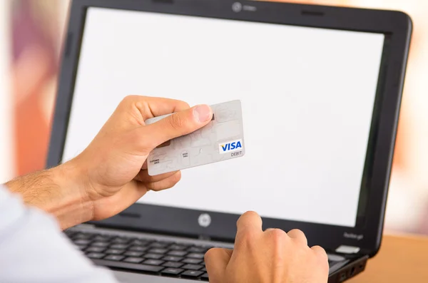Closeup of man sitting by desk with laptop computer holding up Visa credit card in front of screen as in shopping online