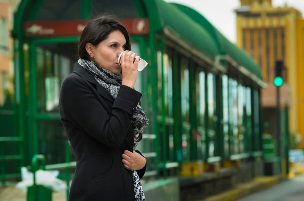 Brunette model wearing black jacket and grey scarf waiting for public transportation acting cold at station with cup of coffee