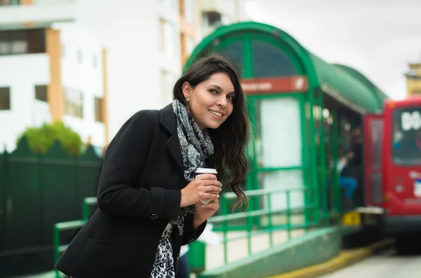 Brunette model wearing black jacket and grey scarf waiting for public transportation acting cold at station with cup of coffee