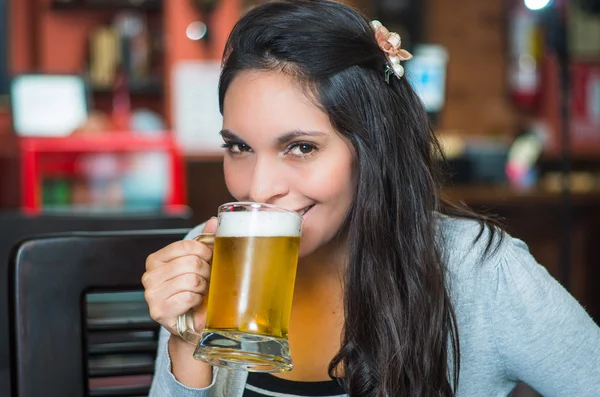Brunette model sitting by restaurant table drinking from glass of beer and posing with positive attitude smiling