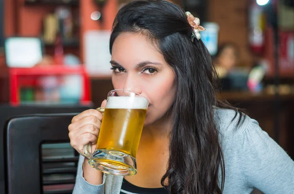 Brunette model sitting by restaurant table drinking from glass of beer and posing with positive attitude smiling