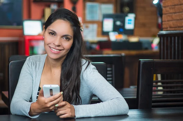Attractive brunette wearing grey sweater sitting by restaurant table holding mobile phone ad smiling to camera