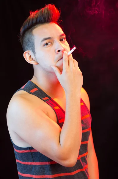 Hispanic male wearing red black striped singlet posing with serious facial expression and smoking a cigarette smoke visible