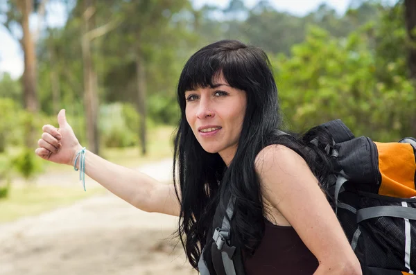 Adventurous brunette, outdoors forest environment wearing backpack, holding thumb out as in hitchiking