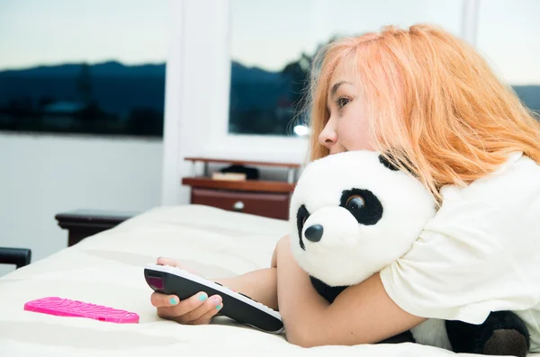 Pretty young woman lying comfortably on bed hugging stuffed panda animal and pressing remote control