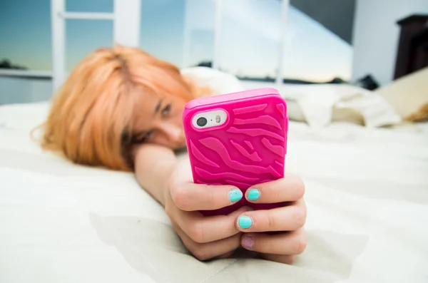Pretty young woman lying comfortably on bed and using pink mobile phone with large windows background