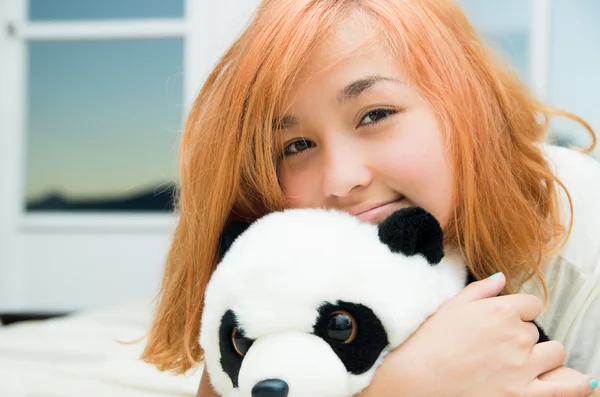 Pretty young woman lying comfortably on white bed and hugging stuffed panda animal with large windows in background