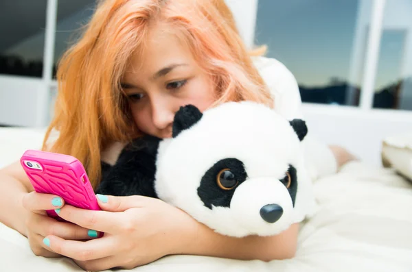 Pretty young woman lying comfortably on bed hugging stuffed panda animal and using pink mobile phone with large windows background