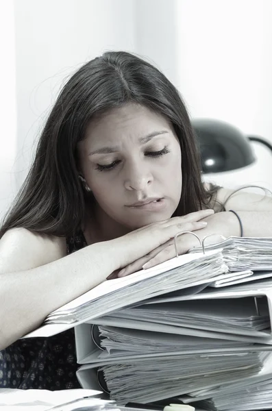 Hispanic brunette office worker sitting by desk and lying over pile of archive files looking tired, black white edition