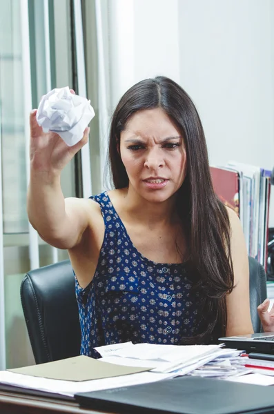 Hispanic brunette sitting by office desk holding ball of paper and upset facial expression