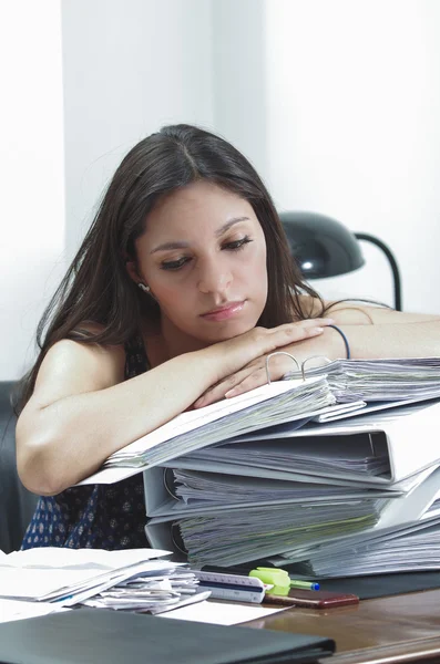 Hispanic brunette office worker sitting by desk and lying over pile of archive files looking tired