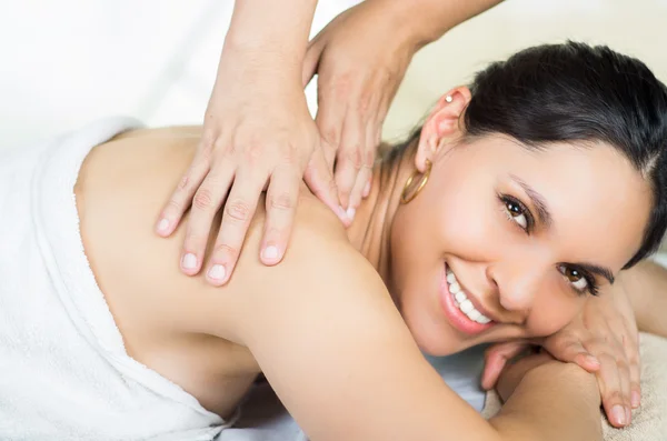 Hispanic brunette model getting massage spa treatment, white towel covering upper body lying horizontal smiling to camera with hands working on shoulders