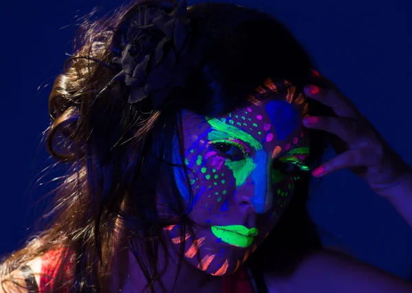 Headshot woman wearing awesome glow in dark facial paint, blue based with other neon colors and obscure abstract background, facing camera