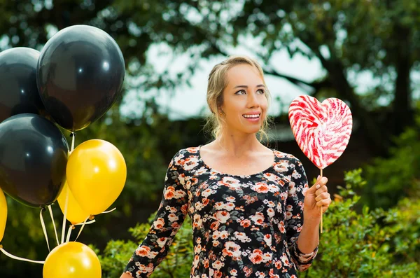 Beautiful model wearing summer dress in garden environment, holding black golden balloons and large lollipop posing happily for camera