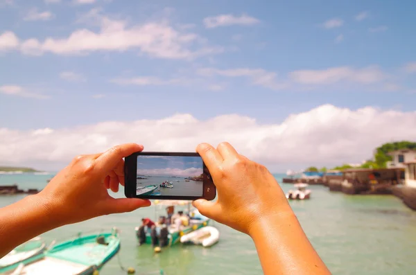 Hands holding mobile phone taking picture of beautiful island bay area on Galapagos islands with nice weather and small boats