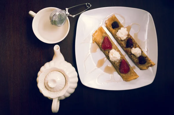 Two beautiful crepes pancakes lying on white plate, decorated with small amount of cream and berries, tea cup next to it, elegant presentation