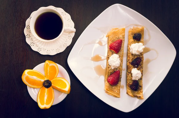 Two beautiful crepes pancakes lying on white plate, decorated with small amount of cream and berries, coffee cup next to it as well sliced orange, elegant presentation