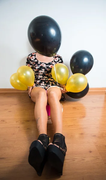 Model wearing summer dress sitting on wooden floor holding black and golden balloons, face covered by balloon