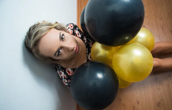 Beautiful model sitting on wooden floor leaning back against white wall peeking head out from black and golden balloons