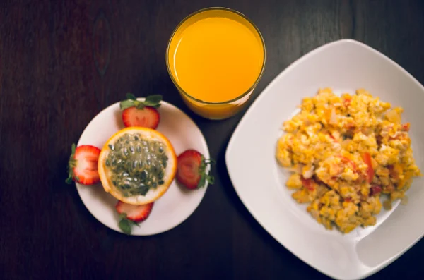 Plate of scrambled eggs, next to it a glass with orange juice and an open genadilla sorrounded by cut strawberries