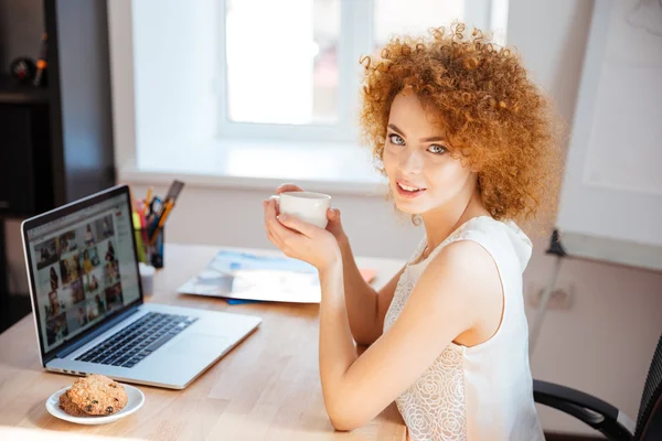 Beautiful smiling woman drinking coffee at table and using laptop