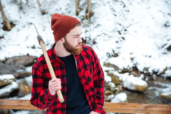 Pensive man in checkered shirt holding axe at winter forest
