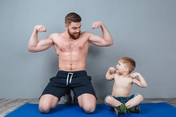 Smiling shirtless dad and son showing biceps sitting on mat