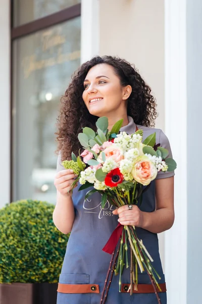 Happy charming young woman florist holding flower bouquet near shop