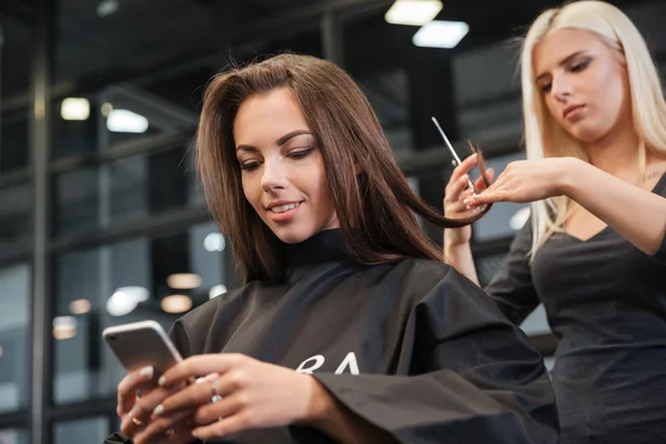 Woman with smartphone and hairdresser making hair styling at salon