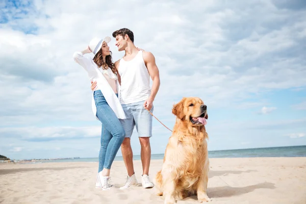 Dog sitting in front of couple standing on the beach