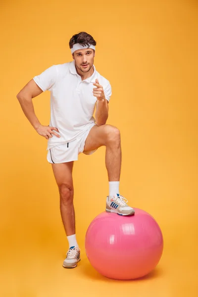 Young handsome sportsman with fitness ball pointing at camera