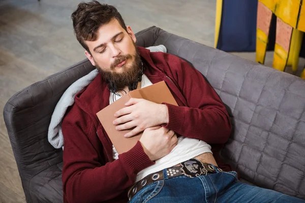 Tired guy with beard sleeping on sofa hugging book