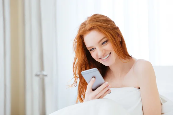 Smiling redhead woman using smartphone in bed