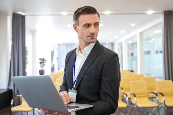 Serious businessman holding laptop in empty meeting hall