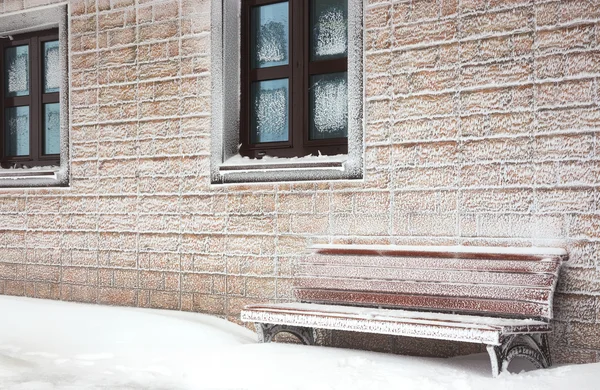 Snow-covered bench at  hoarfrost wall of house