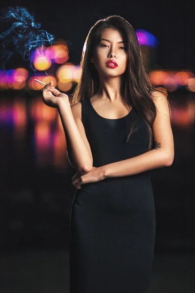 Beautiful asian lady smoking, night city behind her.