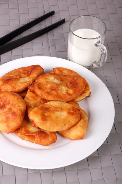 Fried cakes on white plate, kitchen tongs and cup of milk on gra